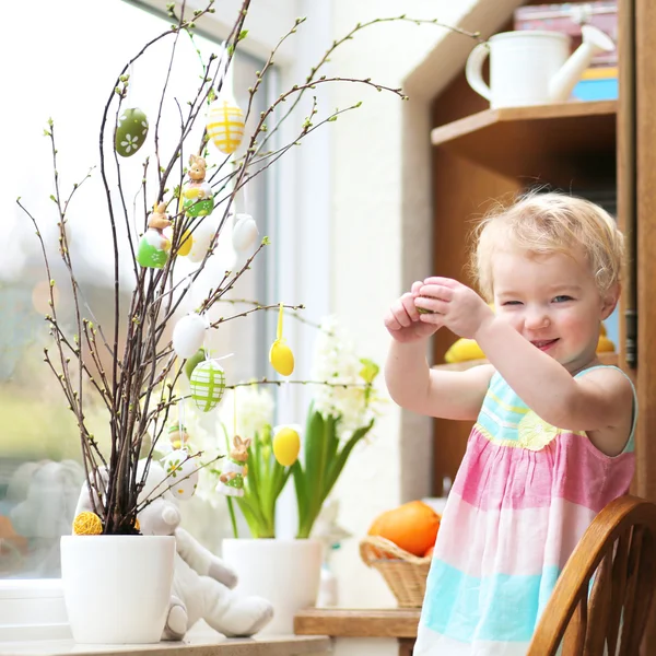 Adorable petite fille blonde tout-petit décorant avec des oeufs de Pâques branches de cerisier debout dans la cuisine à côté d'une fenêtre avec vue sur le jardin — Photo