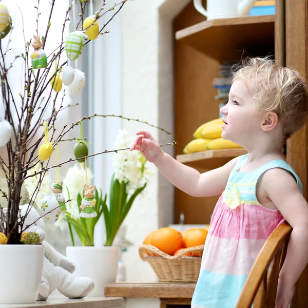 Adorable little blonde toddler girl decorating with Easter eggs cherry tree branches standing in the kitchen next a window with garden view — Stock Photo, Image