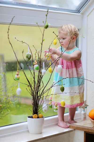 Adorable little blonde toddler girl decorating with Easter eggs cherry tree branches standing in the kitchen next a window with garden view