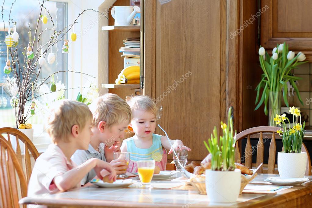 Group of three kids, twin brothers with their little toddler sister, eating eggs during family breakfast on Easter day sitting together in sunny kitchen. Selective focus on little girl.