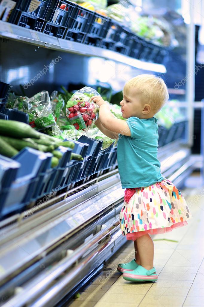Cute baby girl is picking up radish pack from a shelf in vegetables department in a supermarket