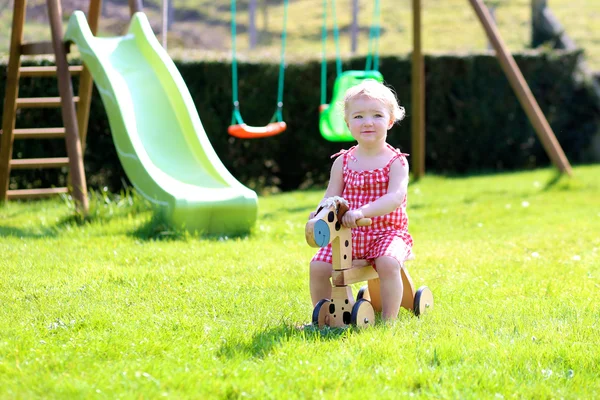 Cute blonde toddler girl riding wooden horse in summer garden — Stock Photo, Image