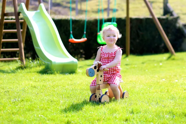 Cute blonde toddler girl riding wooden horse in summer garden — Stock Photo, Image