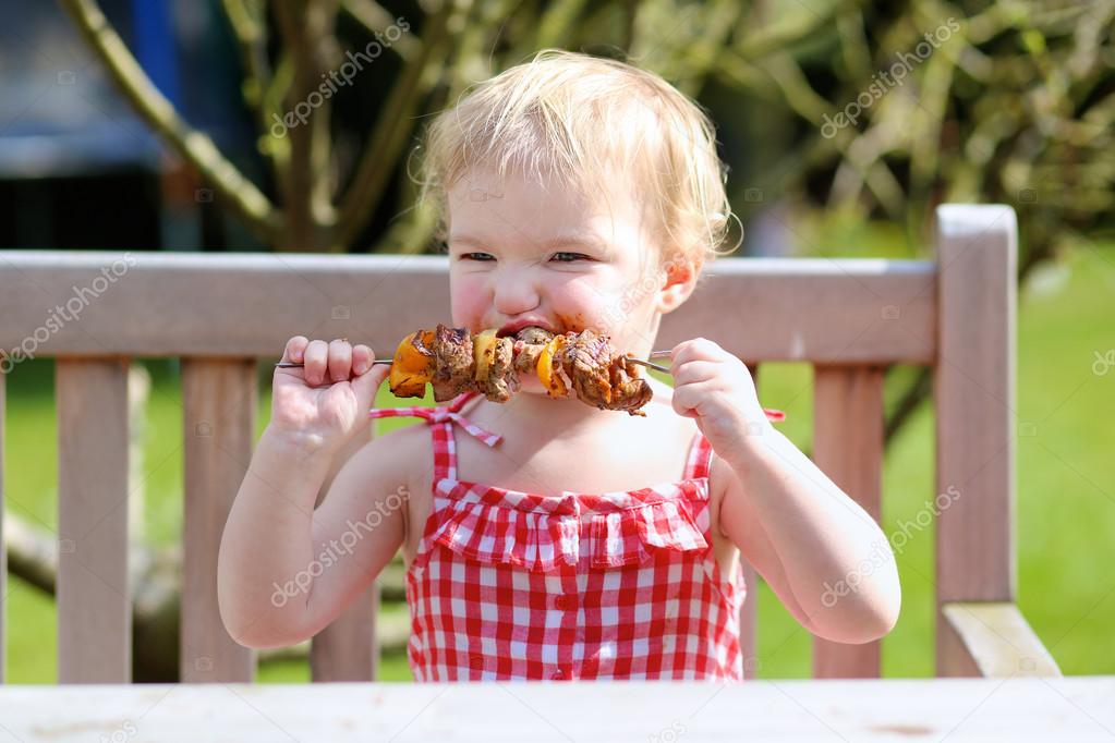 Happy toddler girl eating grilled meat outdoors