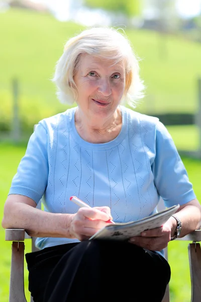 Felice donna anziana rilassante all'aperto seduto in giardino sedia di legno lettura giornale e risolvere enigmi — Foto Stock