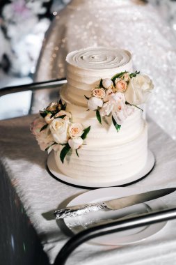 Düğün pastası. İştah açıcı pasta. Bej bir masada üç kat, arka planda süslenmiş güller.