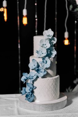 Düğün pastası. Şekerler. Düğün pastası siyah zemin üzerinde üç kat beyaz mastik süslemeli düğün günü mastikten mavi ve mavi orkidelerle süslenmiş.