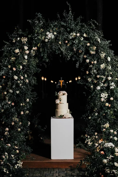 Düğün pastası. Bej bir masada üç katlı pasta, bir düğün kemerine karşı güllerle süslenmiş.