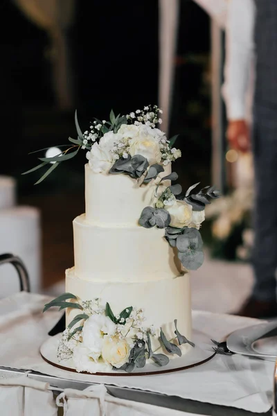 A wedding cake. Appetizing cake three tiers on a beige table, decorated roses, eucalyptus, gold leaf on a dark background