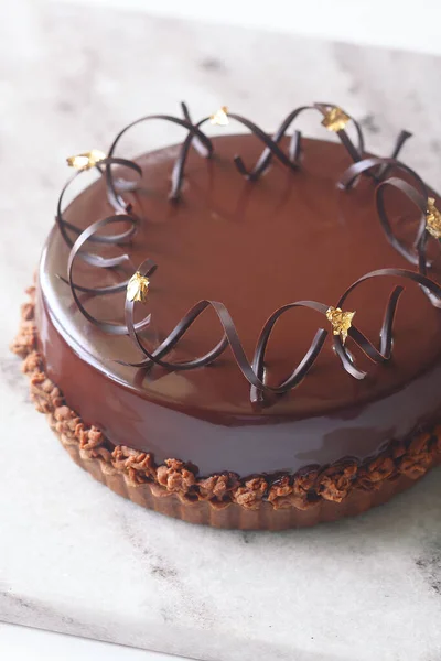 Contemporary Chocolate Praline Mousse Cake, covered with chocolate mirror glaze and garnished with chocolate spirals, on light gray marble board.