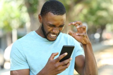 Siyah tenli kızgın adam parkta akıllı telefon içeriğini kontrol ediyor.
