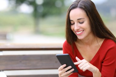 Parktaki bankta oturan mutlu bir kadın akıllı telefon içeriğini araştırıyor.