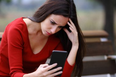 Worried woman sitting on a bench checking smart phone content in a park