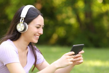 Kulaklıklı Asyalı kadın yeşil bir parkta akıllı telefondan video izliyor.
