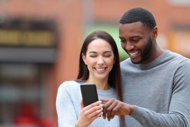 Front view portrait of a happy interracial couple consulting phone text in the street