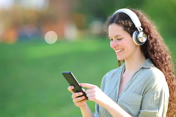 公園でヘッドフォンを身に着けている音楽を聞いて幸せな女性はスマートフォンをチェック — ストック写真