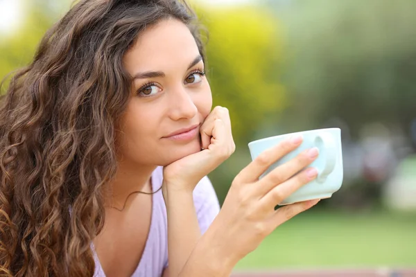 自信的女人看着坐在公园里拿着咖啡杯的相机 — 图库照片