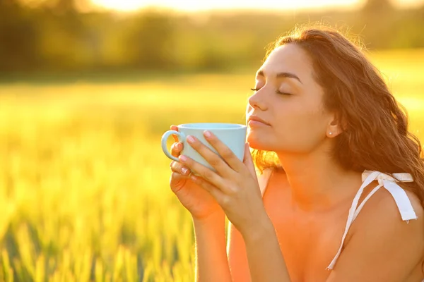 Woman relaxing drinking coffee sitting in a wheat field at sunset