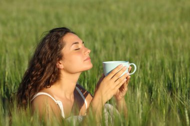 Woman relaxing smelling coffee from cup in a green wheat field