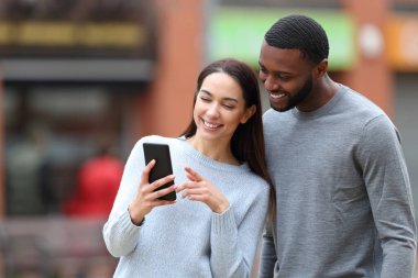 Happy interracial couple checking cell phone in the street