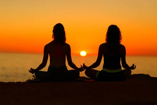 Back view backlight portrait of two women silhouettes doing yoga at sunset on the beach