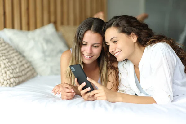 Two happy friends checking smartphone lying on a bed
