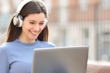 Kulaklıklı dizüstü bilgisayarı olan mutlu bir kadın sokakta bankta oturuyor.