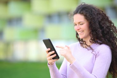 Kıvırcık saçlı mutlu kadın sokakta akıllı telefon içeriğini kontrol ediyor.