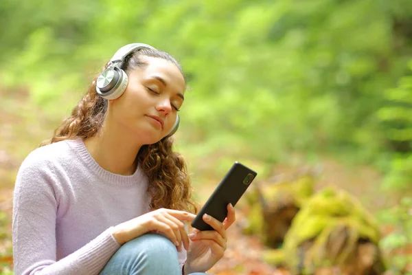 美丽的女人坐在森林里 一边听音乐 一边带着耳机 一边放松地听着电话 — 图库照片