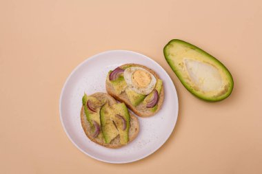 Avokado ile iki sandviç, haşlanmış yumurta ve beyaz tabakta kırmızı soğan, yarısı bej arka planda avokado. Asgari biçim kompozisyonu.