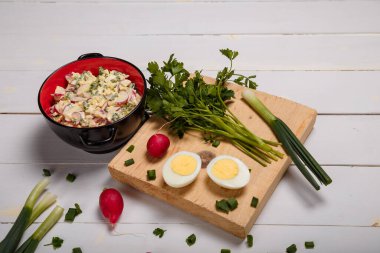 Turp, yeşil soğan, maydanoz, kırmızı kasede yumurta salatası beyaz ahşap arka planda mutfak tahtası. Sağlıklı yeme alışkanlıkları, organik gıda, diyet ve beslenme konsepti.