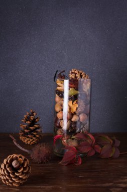 Çam kozalaklı şeffaf cam vazo, fındık, kestane, kuru yapraklar, meşe palamudu, koyu arkaplanda kalça gülü böğürtlen. Sonbahar kompozisyonu, dikey çekim