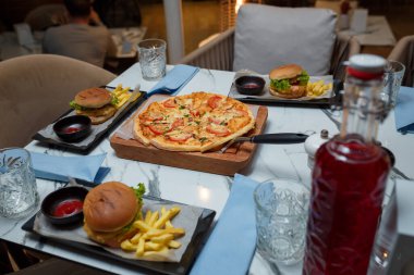 Tahtada domates ve peynirli pizza, hamburger ve patates kızartması, içecek bardakları. Restoran masasında şölen yemeği.
