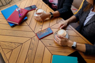 İki meslektaş iş yemeği yiyor, kahve molası. Elleri kapuçino bardaklarını tutuyor. Defterler, akıllı telefonlar, kalemler ve gözlükler ahşap masaya.