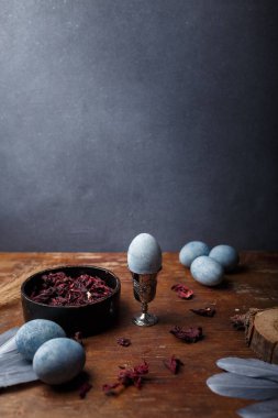 Mavi boyanmış paskalya yumurtaları, yumurta fincanı, kuru Hibiscus çayı ve ahşap arka planda gümüş dekoratif tüyler, kopya alanı, dikey çekim alanı