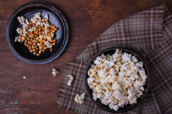 Heap of ready and raw popcorn in black bowl on wooden rustic background. Some popcorns on fabric and table