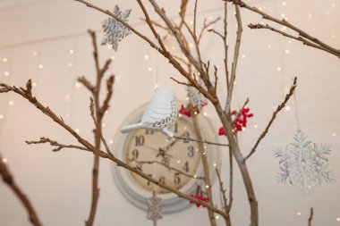 Kuru ağaç dalında asılı beyaz kuş oyuncağı. Kar taneleri, kırmızı böğürtlenler ve altın çelenk ışıkları...