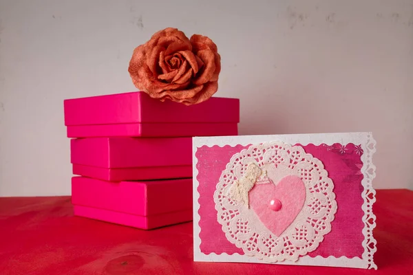 Three bright pink present boxes, fabric rose flower, postcard with heart on painted wooden background. Happy Valentines day