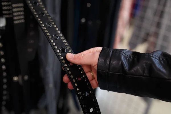 Male hand in leather jacket sleeve holding a belt with metallic rivets. Choosing accessory in department store or supermarket, modern fashion