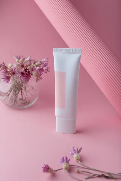 Tender cosmetics tube on pink paper background with flowers, mockup no brand template, copy space. Face skin care with natural extract