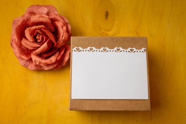 Present box with blank white postcard, fabric rose flower on bright yellow background, top view. Birthday, wedding, mothers, Valentines day
