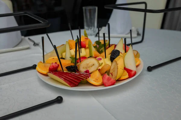 Festive fruits plate with cut fruit pieces on skewers in white plate. Dessert for birthday party, sweet appetizer. Banana, apple, pear and prunes, orange, strawberry and mint