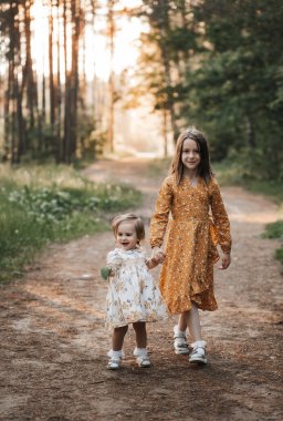 İki kız kardeş el ele tutuşarak ormanda koşuyor. Yaz güneşli bir gün ve açık elbiseli kızlar. 