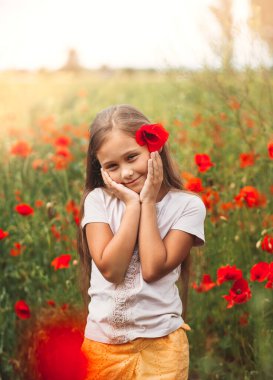 Yaz güneşinde haşhaş tarlasında poz veren uzun saçlı küçük kız. Dikey