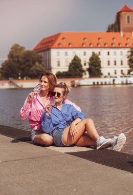 Smiling best friends girls drink wine on a walk. Portrait of two happy girls romantically sitting on rivwr background with bottle in hand, talking and smiling. Emotions concept.