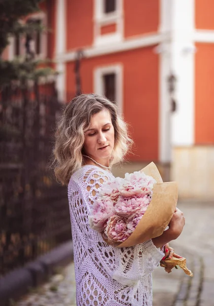 Positive girl hold flowers  in the city center. Close up shot of amazing blonde haired lady with natural make up look happy, walking outdoors  hold pink bouquet.