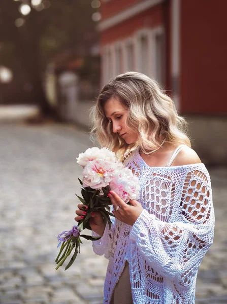 Positive girl hold flowers  in the city center. Close up shot of amazing blonde haired lady with natural make up look happy, walking outdoors  hold pink bouquet.
