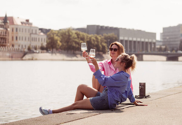 Smiling best friends girls drink wine on a walk. Portrait of two happy girls romantically sitting on rivwr background with bottle in hand, talking and smiling. Emotions concept.