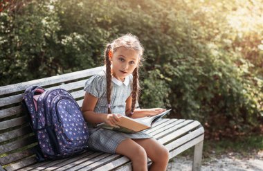 Girl with a backpack sitting on a bench and holds  books near the school. Back to school, lesson schedule, a diary with grades. Education, primary school classes, Back to school