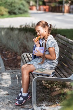 Back to school. Cute little school girl sitting on bench in school yard and eating lunch outdoor. Right school meal for lunch